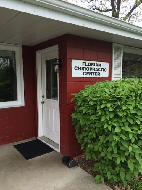 The front door of Florian Chiropractic: a white door beside a white sign reading “Florian Chiropractic Center” on the red wall, with a leafy shrub.