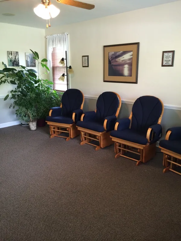 A row of navy upholstered glider rocking chairs in the waiting room, with a large potted plant and framed pictures.