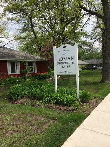 The white post sign on the lawn at 557 Greenleaf Street reading “557 Florian Chiropractic Center”, with lilies around its base and the red office behind it.