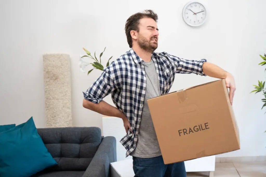 A man holding his lower back while carrying a cardboard box marked “Fragile”.